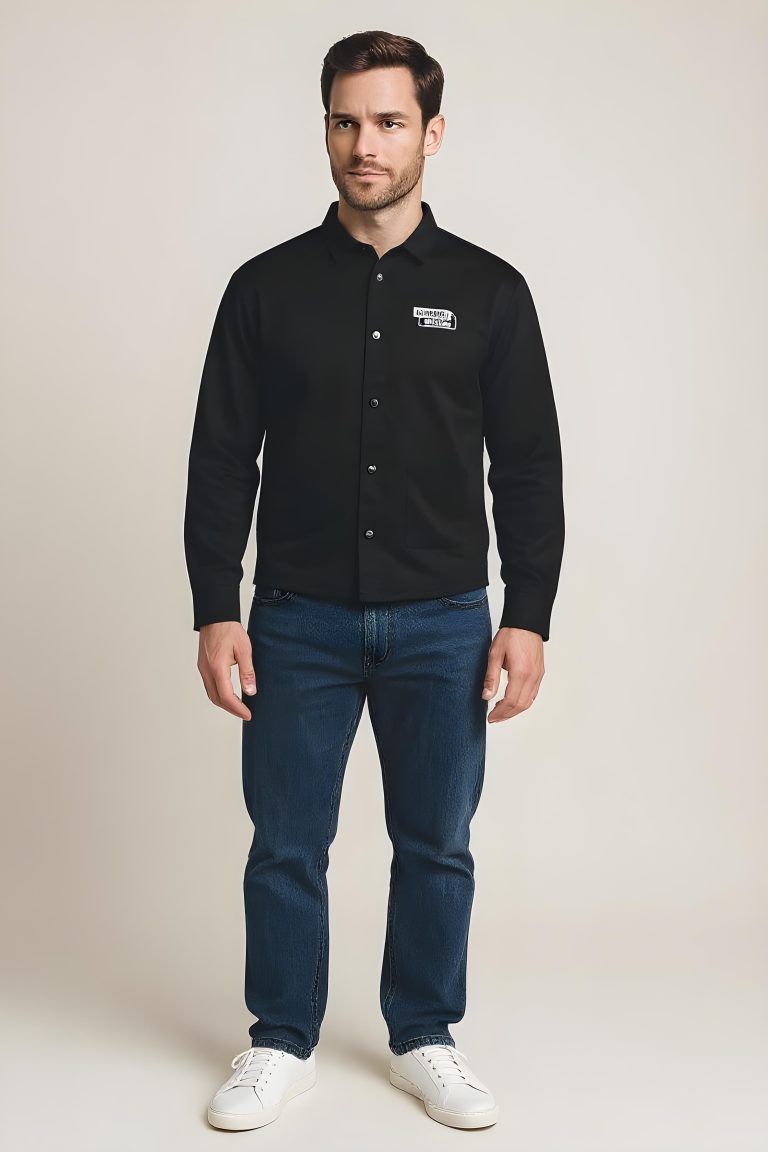 A man stands facing forward, wearing a black button-up shirt with a name patch, blue jeans, and white sneakers against a plain light background.