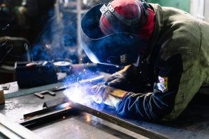 A person wearing protective gear welds metal pieces together on a workbench, with sparks and blue light visible from the welding process.