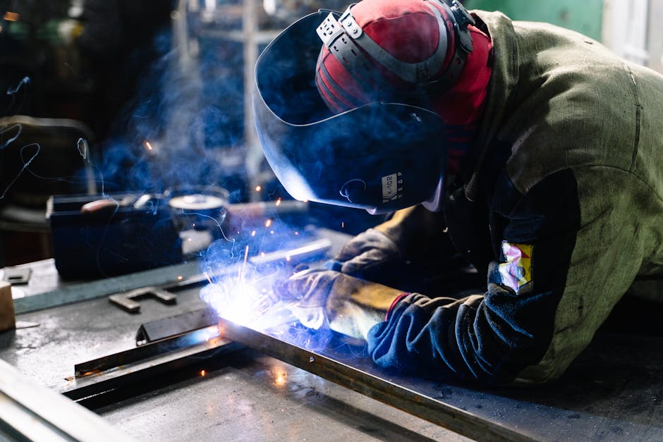 A person wearing protective gear welds metal pieces together on a workbench, with sparks and blue light visible from the welding process.
