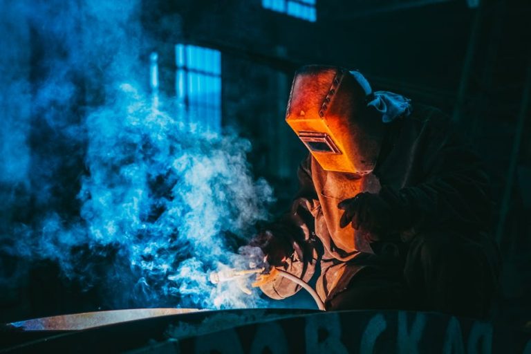 A person wearing a protective welding helmet and gear works with welding equipment, producing bright sparks and blue smoke in an industrial setting.