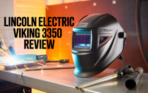 A Lincoln Electric Viking 3350 welding helmet sits on a metal table with welding tools and sparks in the background. Text reads: "Lincoln Electric K3034 Review: VIKING 3350 Welding Helmet.